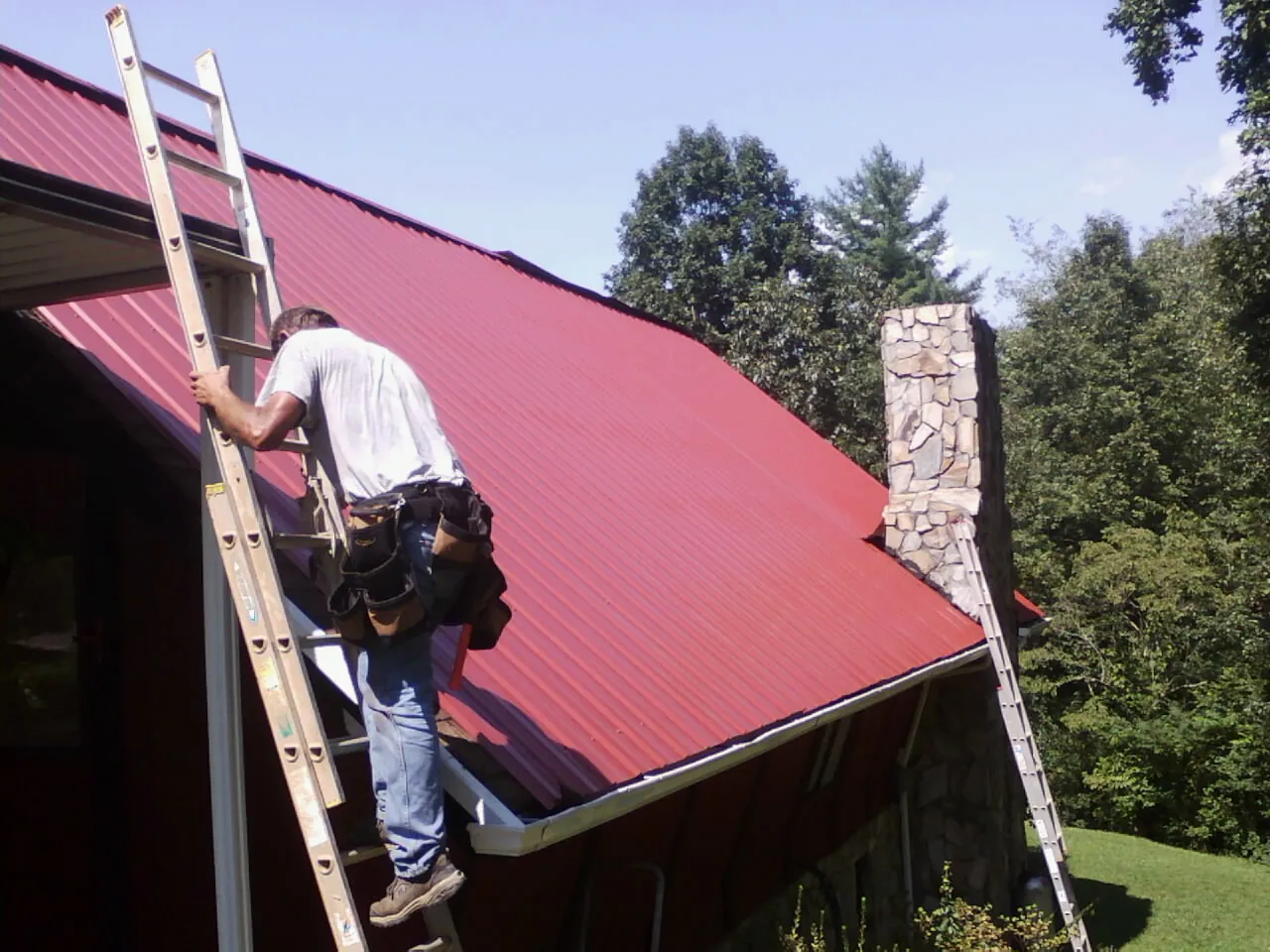 Roofer on a ladder beside a red metal roof and stone chimney — Soffit & Fascia Repair by Roofing Brothers in Litchfield